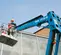 Rear angled view of a blue Genie boom lift with a worker in the basket operating the control panel on a building facade.