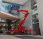Rear view of a red atrium lift with extended arm and stabilizers, operator using lift indoors near glass windows.
