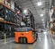 Rear view of an orange and grey electric Toyota warehouse forklift in a warehouse isle with an operator in an open cabin.