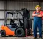 Side view of an orange and grey Toyota warehouse forklift with a black mast in a building with a worker standing beside it.