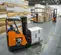 Side view of an orange and black Toyota electric warehouse forklift with an operator lifting boxes in an indoor warehouse.