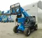 Front angled view of a blue Genie telehandler with extended boom and fork attachment with open cabin on a construction site.