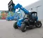 Front angled view of a blue Genie telehandler with extended boom and fork attachment with open cabin on a construction site.