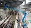 Side view of blue boom lift in indoor warehouse, extended over pipes, operator using lift for maintenance, for rent.