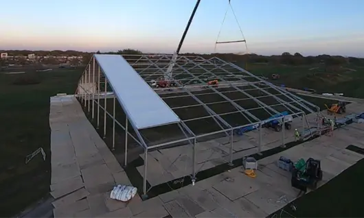 A crane installing a roof panel on a large temporary fabric structure at dusk in an open field
